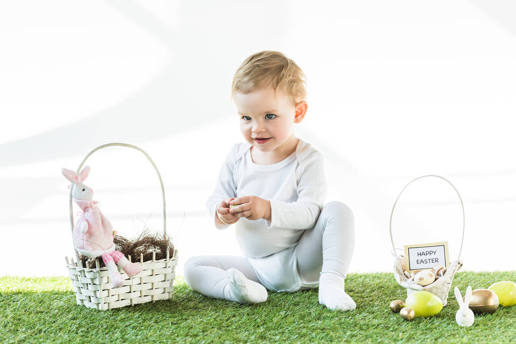 cheerful baby sitting near straw baskets with Easter eggs, decorative rabbits and happy Easter card isolated on white - Photo, Image