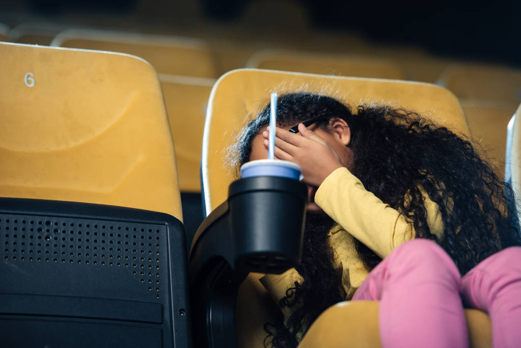 pelästynyt afrikkalainen amerikkalainen lapsi kädestä kasvoilla istuessaan elokuvateatterin istuimella paperimuki kupin pidikkeessä
 - Valokuva, kuva