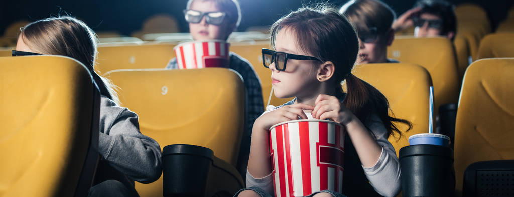 панорамний знімок дітей, які дивляться фільм і тримають паперові чашки з попкорном
 - Фото, зображення