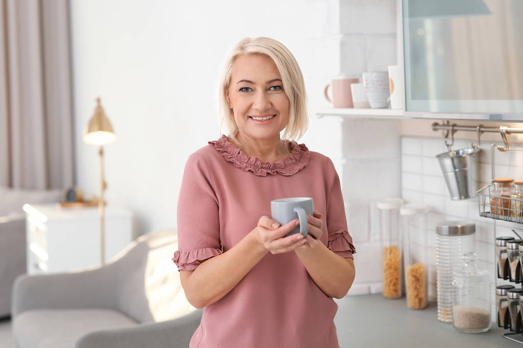 Portret van mooie rijpe vrouw met cup in keuken - Foto, afbeelding
