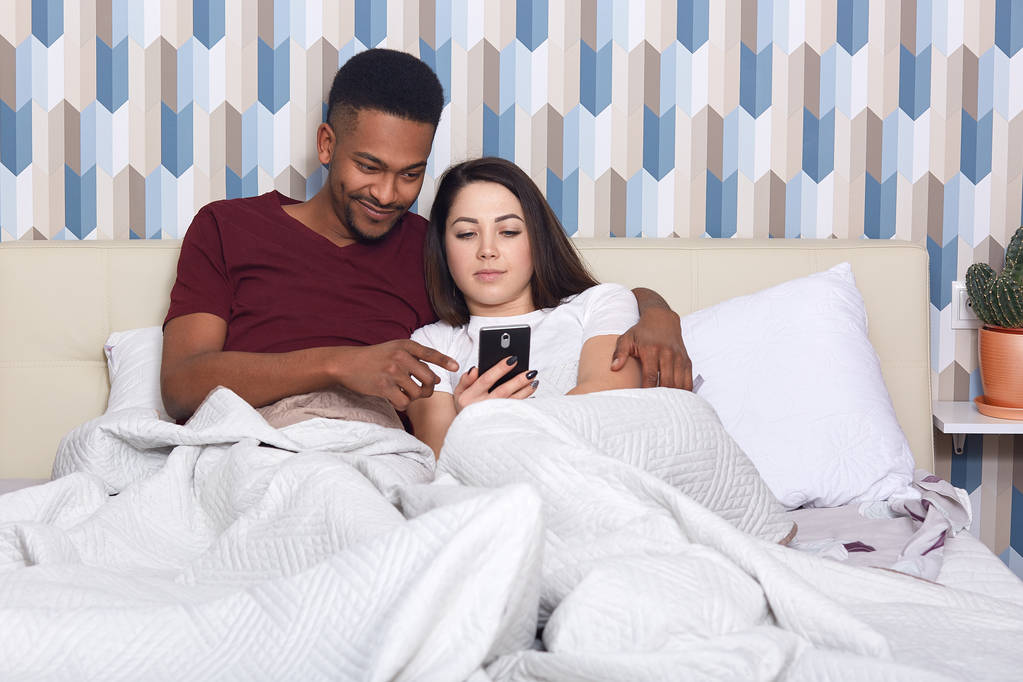 Young couple lying in bed, female iholding mobile phone, husband hugs his wife, watching film online, having rest together, isolated on colourful wall. People, relation, resting, comunication concept. - Photo, Image