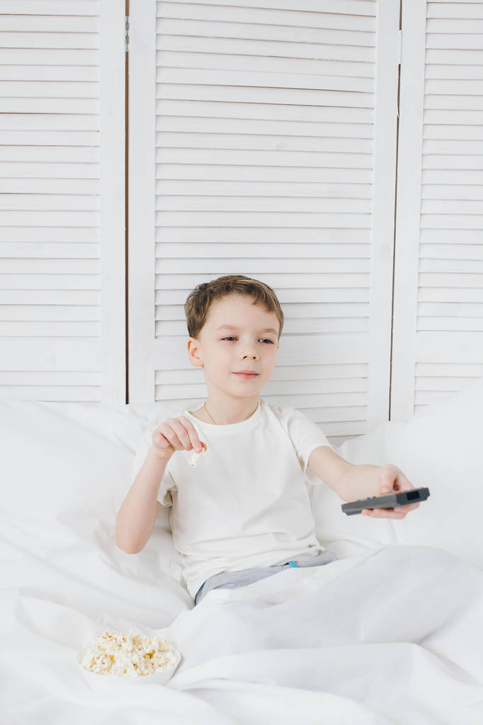 Хлопчик їсть попкорн сидить у ліжку і дивиться телевізор
  - Фото, зображення