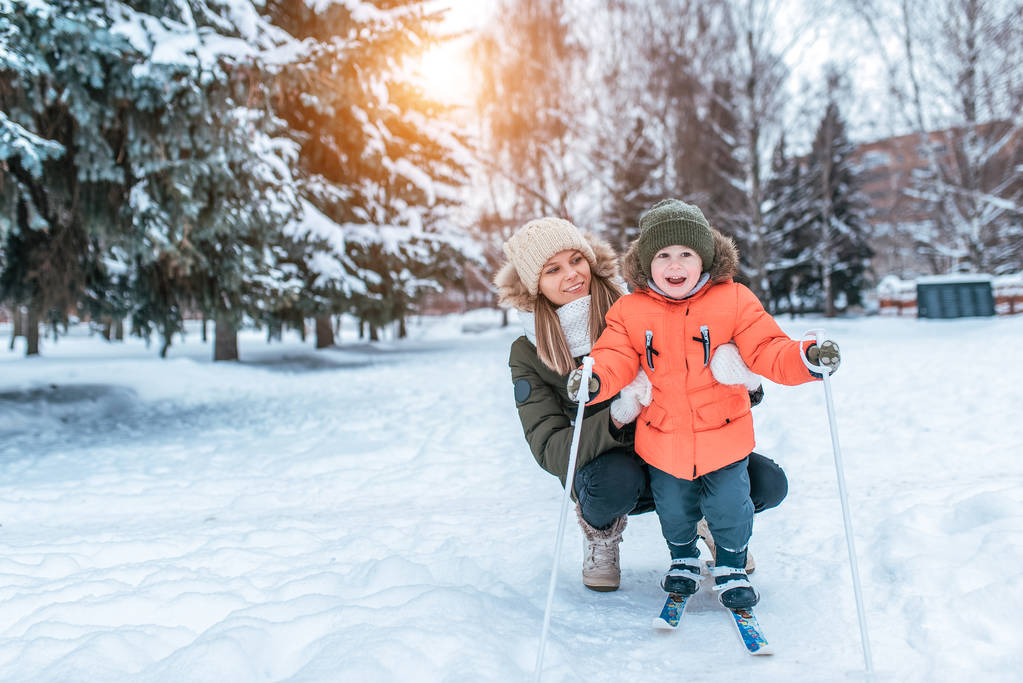 Kobieta matka pomaga jej dziecko, chłopiec 3-6 lat, stoisko dla dzieci narty, radość uśmiech zabawa. Pierwsze kroki na nartach. Wsparcie, uczenie się zdrowego trybu życia. W zimie, w parku miejskiego lasu. Ilość wolnego miejsca dla tekstu. - Zdjęcie, obraz