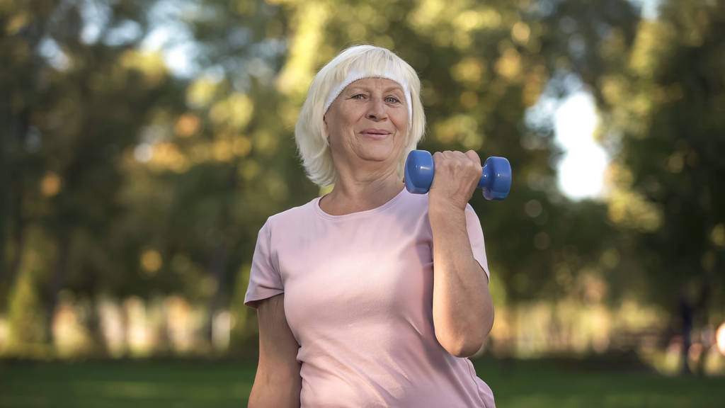 Dame aînée souriante faisant du fitness avec des haltères lors d'une journée ensoleillée dans le parc, mode de vie
 - Photo, image