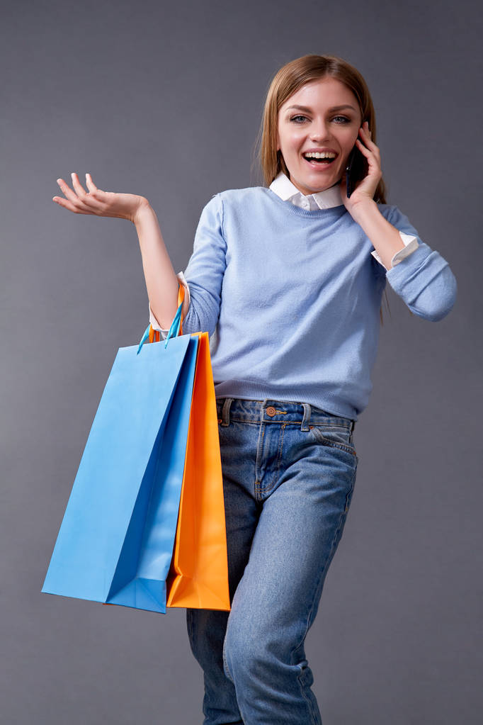 Молодая женщина разговаривает на вашем смартфоне стоя с цветными сумками для покупок
. - Фото, изображение