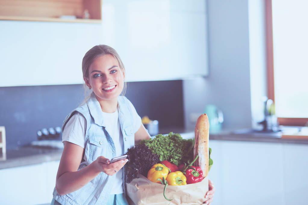Mujer joven sosteniendo bolsa de la compra de comestibles con verduras. De pie en la cocina - Foto, imagen
