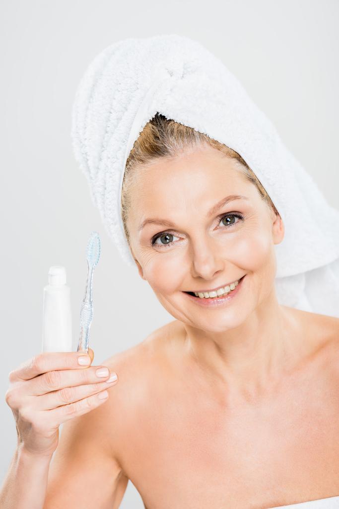 όμορφη και χαμογελαστή ώριμη γυναίκα σε πετσέτα κρατώντας οδοντόκρεμα και οδοντόβουρτσα που απομονώνονται σε γκρι  - Φωτογραφία, εικόνα