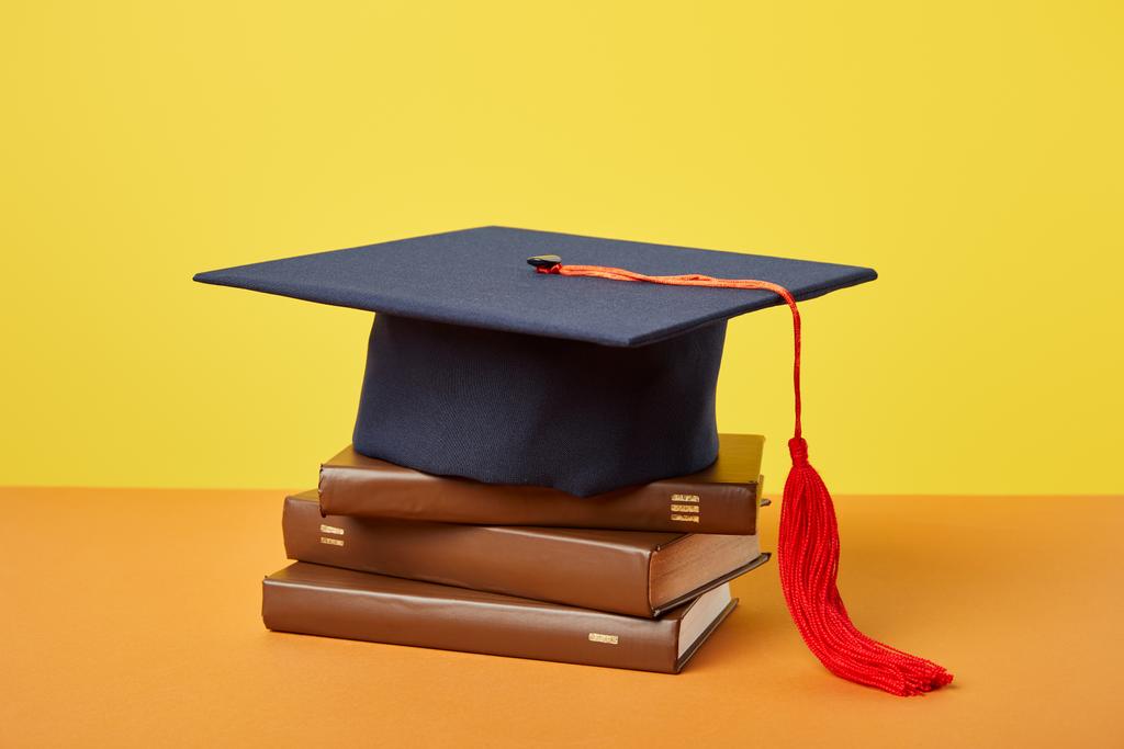 Casquette académique et livres bruns sur surface orange isolés sur jaune
 - Photo, image