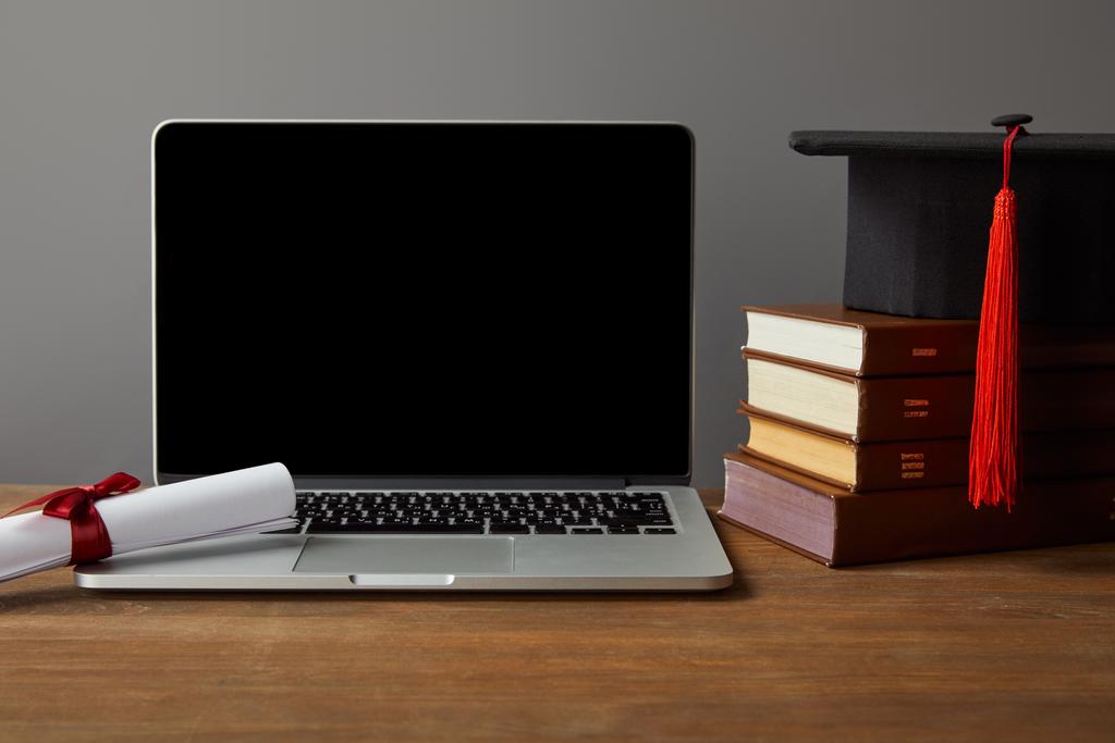 Laptop con schermo bianco, libri, diploma e cappuccio accademico su tavolo in legno isolato su grigio
 - Foto, immagini
