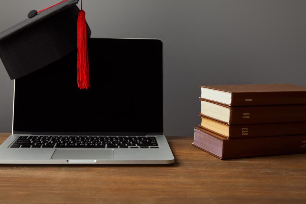 Laptop mit leerem Bildschirm, Bücher und akademische Mütze mit roter Quaste auf hölzerner Oberfläche isoliert auf grau - Foto, Bild
