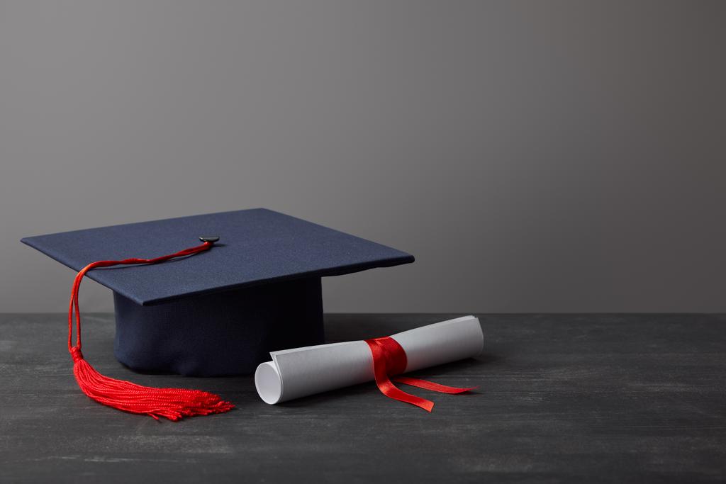 Diploma and academic cap with red tassel on dark surface on grey - Photo, Image