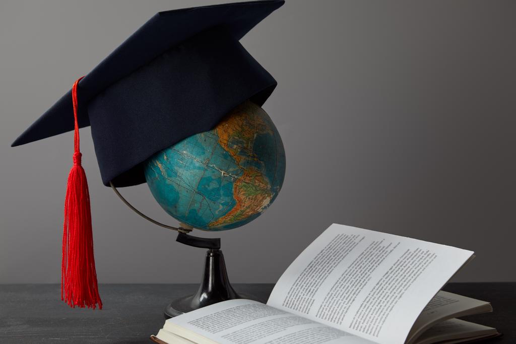 Globe, czapka akademicka z czerwonym pomponem i otwarta książka na powierzchni tekstury na szarym - Zdjęcie, obraz
