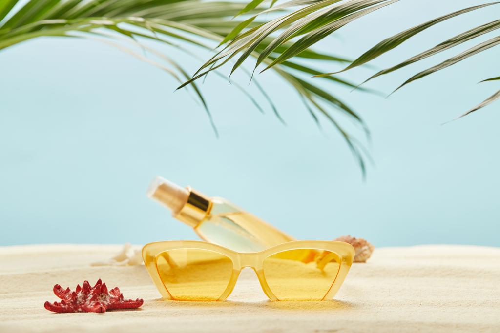 зеленые пальмовые листья рядом с красной морской звездой, желтые солнцезащитные очки и бутылка с маслом загара на песке изолированы на синий
 - Фото, изображение