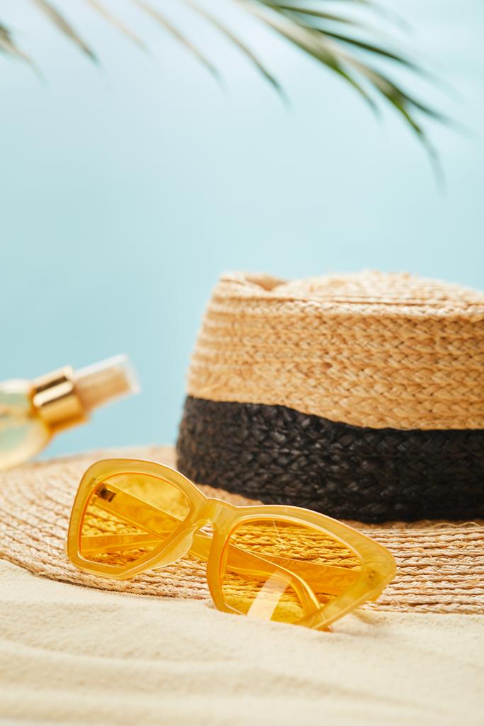 selective focus of sunglasses near straw hat and bottle with suntan oil on sandy beach isolated on blue - Photo, Image