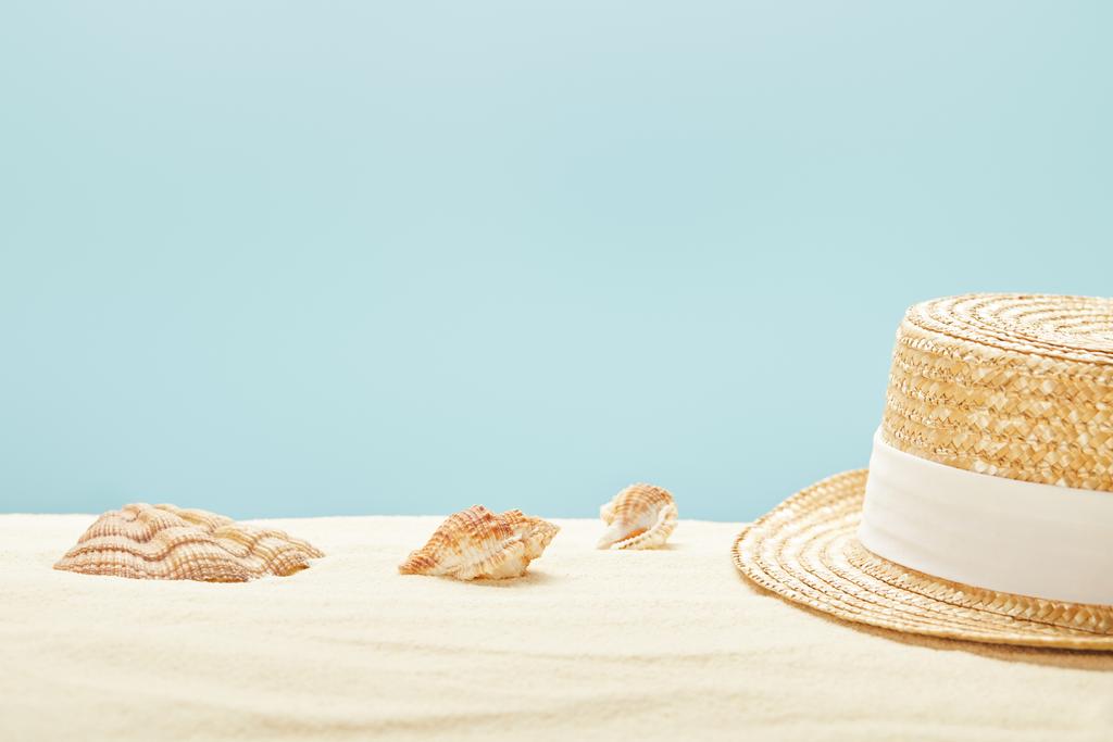 selective focus of straw hat near seashells on sandy beach in summertime isolated on blue - Photo, Image