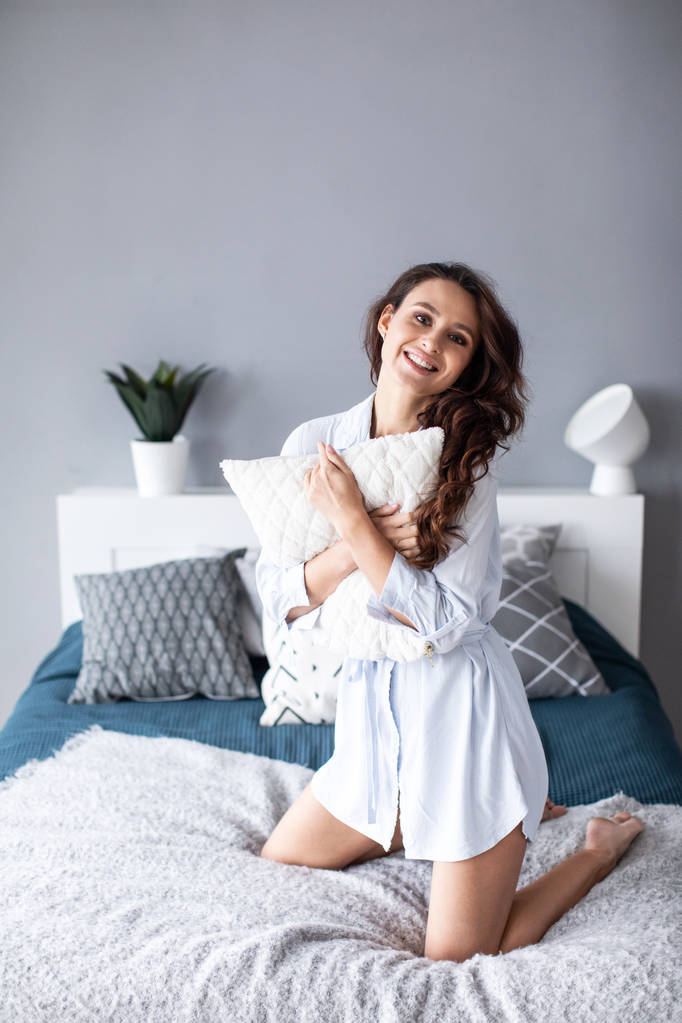 Pozytywna kobieta z poduszką siedzi w łóżku i uśmiecha się do kamery w domu.  - Zdjęcie, obraz
