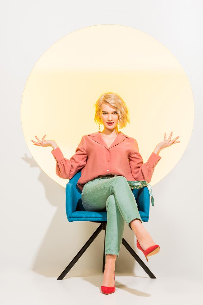beautiful stylish girl in colorful clothes sitting in armchair and gesturing with hands on white with yellow circle - Photo, Image