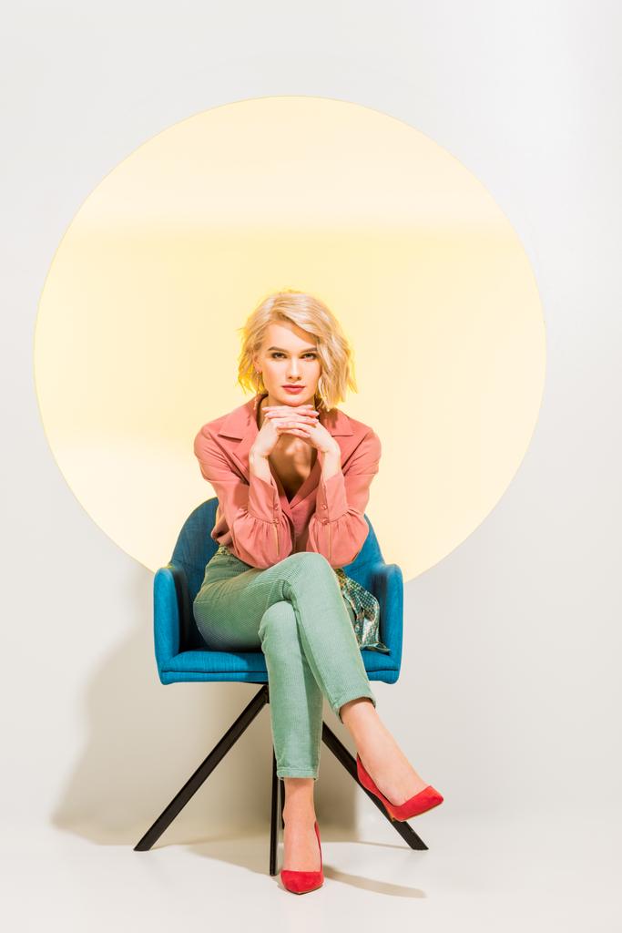 hermosa chica elegante en ropa colorida mirando a la cámara y sentado en sillón en blanco con círculo amarillo
 - Foto, Imagen