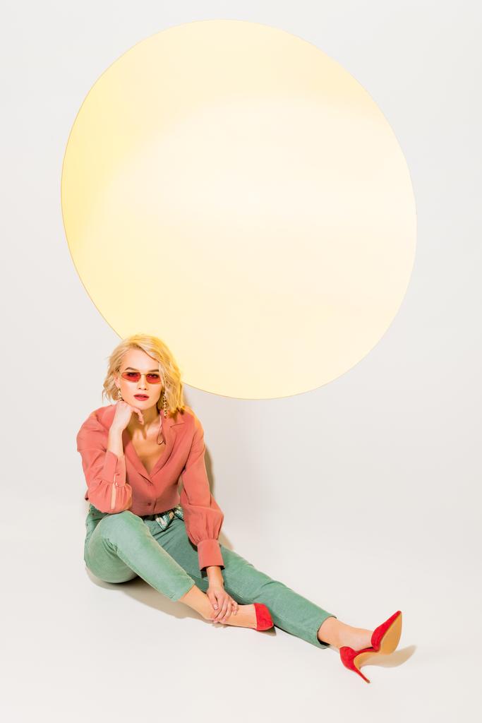 όμορφο κομψό κορίτσι με πολύχρωμα ρούχα και γυαλιά ηλίου που θέτουν σε λευκό με κίτρινο κύκλο  - Φωτογραφία, εικόνα