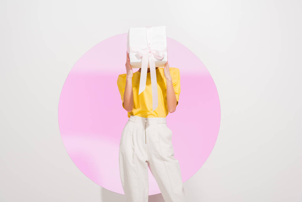 stijlvol meisje Holding bekleding gezicht met geschenkdoos op wit met roze cirkel - Foto, afbeelding