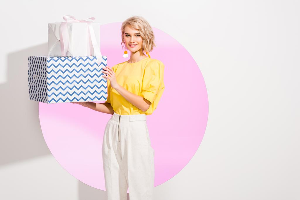 hermosa chica de moda sosteniendo cajas de regalo y sonriendo en blanco con círculo rosa
 - Foto, imagen