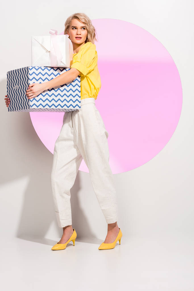 όμορφη μοντέρνα κορίτσι κρατώντας κουτιά δώρων και ποζάρει στο λευκό με ροζ κύκλο - Φωτογραφία, εικόνα