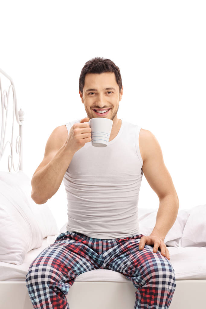 Nuori mies pyjamassa istuu sängyllä ja juo kupin kahvia eristyksissä valkoisella pohjalla
 - Valokuva, kuva