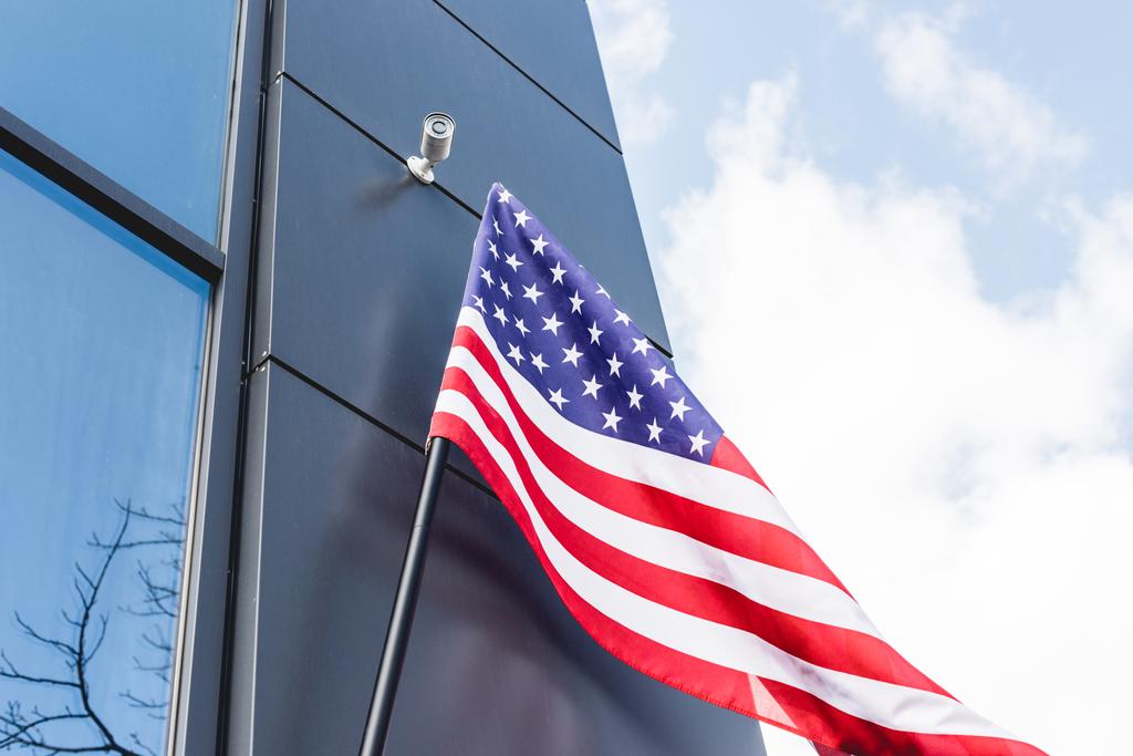 Niedrigwinkel-Ansicht der amerikanischen Nationalflagge mit Sternen und Streifen in der Nähe von Gebäuden gegen blauen Himmel mit Wolken  - Foto, Bild