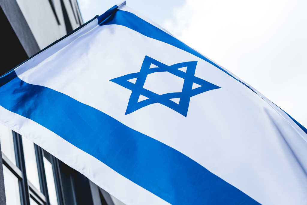 низкоугольный вид национального флага Израиля со звездой Дэвида рядом со зданием против неба
 - Фото, изображение