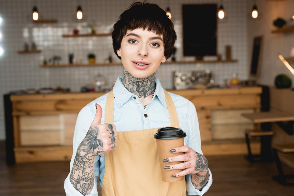 garçonete sorridente em avental mostrando polegar para cima e segurando copo de papel
 - Foto, Imagem
