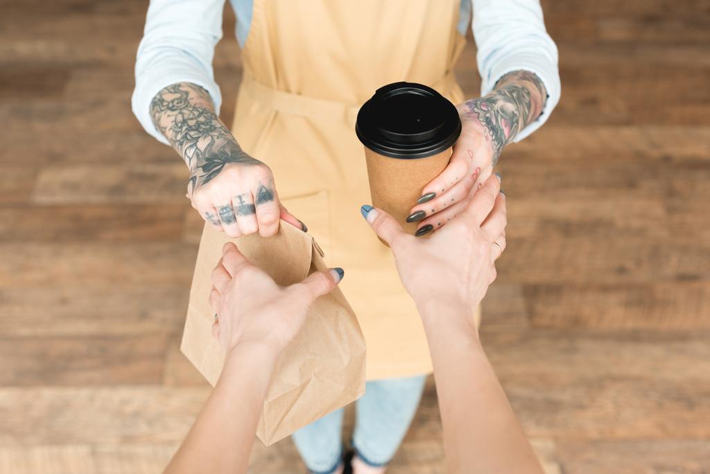 vista recortada de camarera tatuada dando bolsa de papel y taza desechable al cliente
 - Foto, Imagen