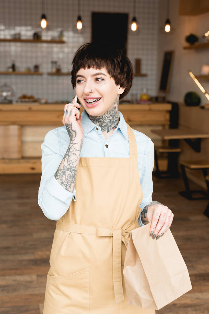 Lächelnde Kellnerin in Schürze spricht mit Smartphone und hält Papiertüte in der Hand - Foto, Bild