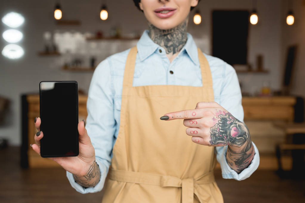 Ausgeschnittene Ansicht der Kellnerin in Schürze, die mit dem Finger auf Smartphone mit leerem Bildschirm zeigt - Foto, Bild
