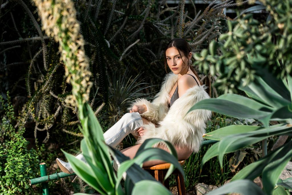 Sexy young woman in white faux fur coat sitting on chair in botanical garden - Photo, Image