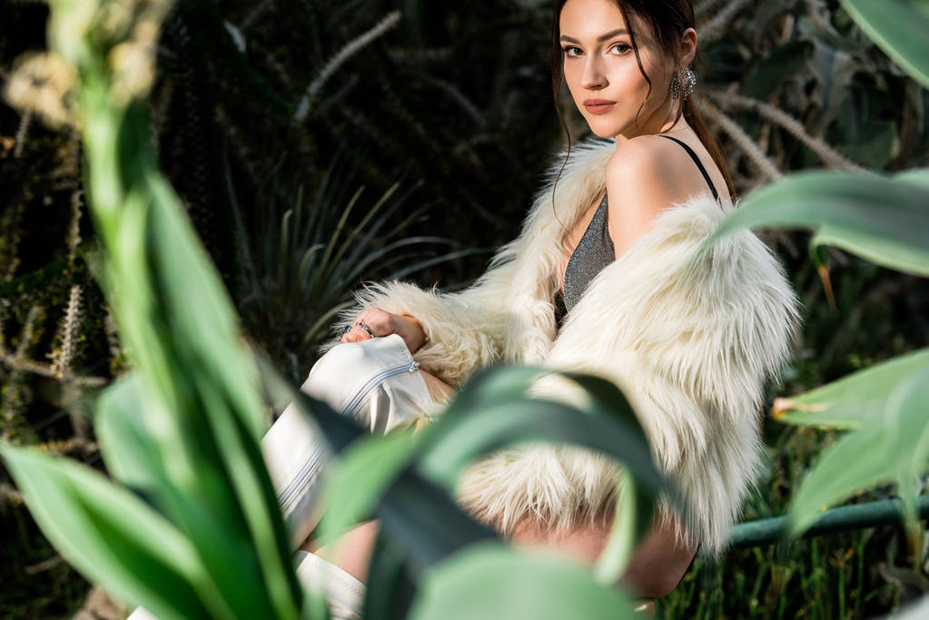 Mujer joven atractiva en abrigo de piel sintética blanca y ropa interior cerca de plantas en jardín botánico
 - Foto, Imagen