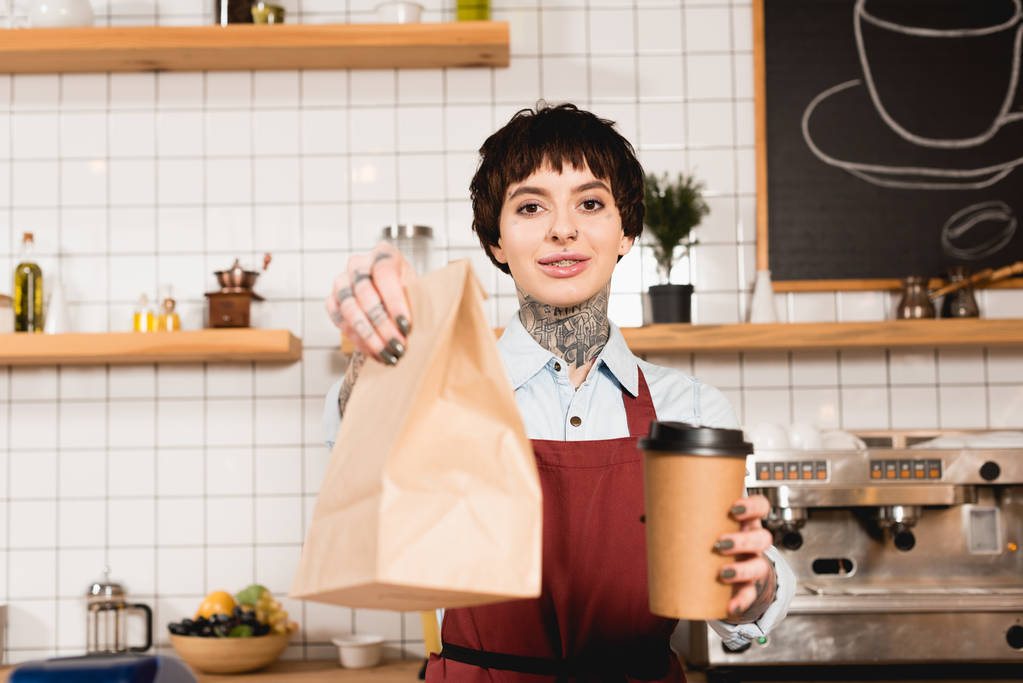 selective focus of smiling barista in apron holding paper bag and disposable cup - Photo, Image