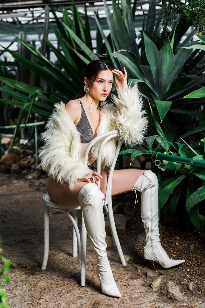 Sexy woman in faux fur coat and white boots sitting on chair in orangery - Photo, Image