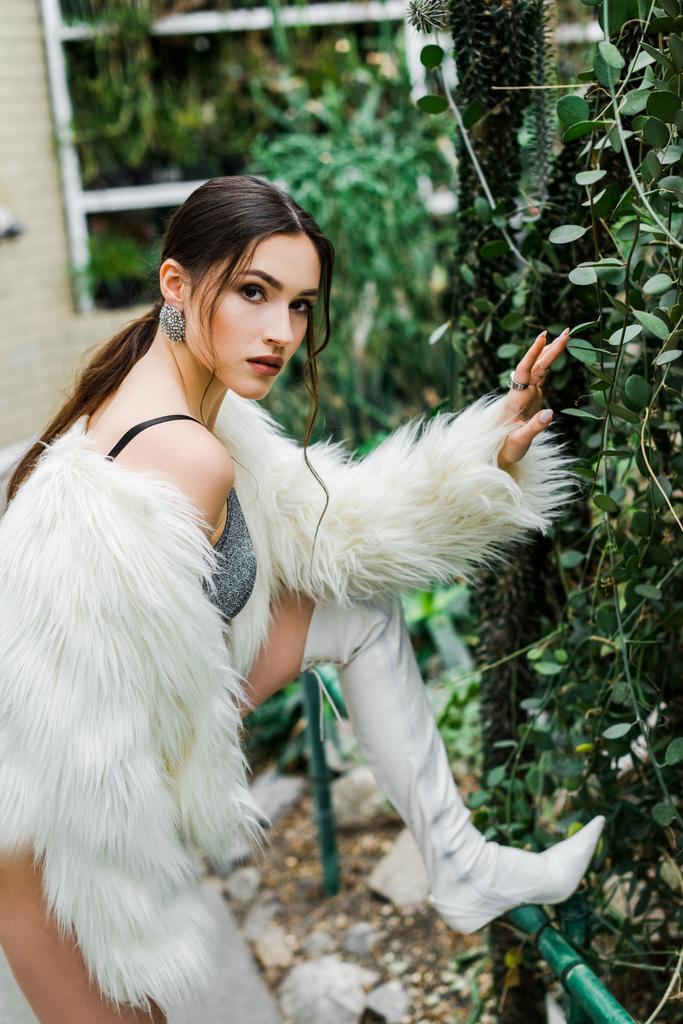 Confident sexy girl in faux fur coat touching green foliage in orangery - Photo, Image
