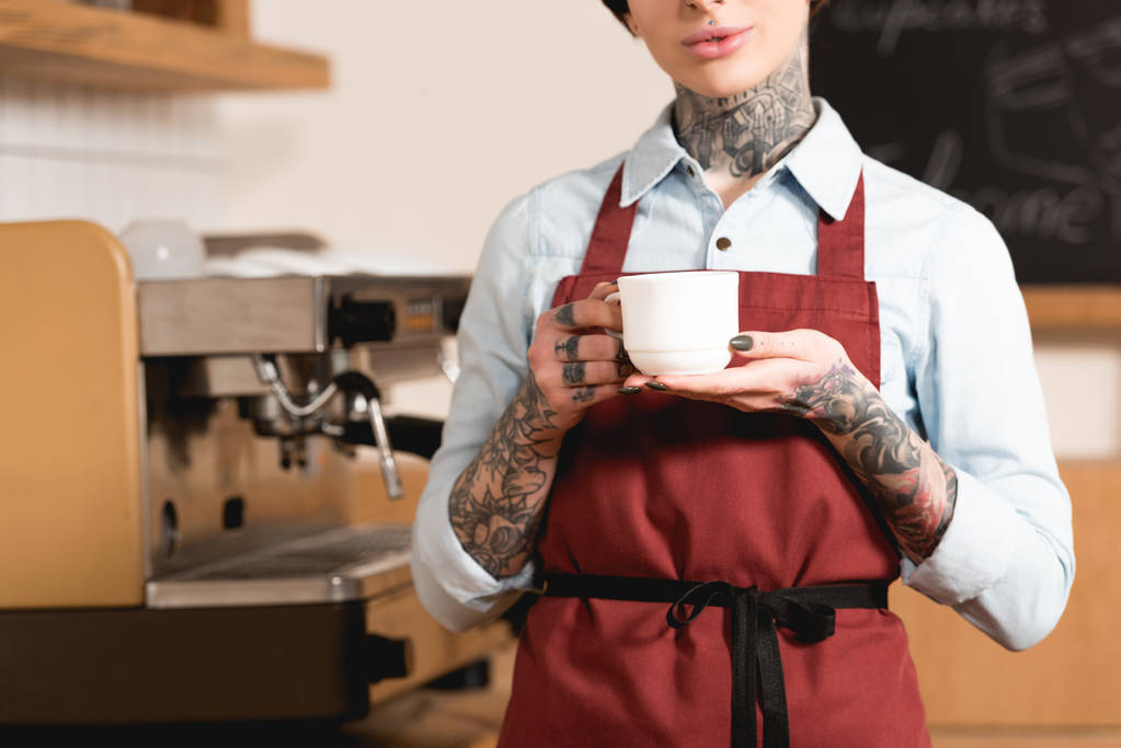 vista parcial de barista em avental segurando xícara de café enquanto está em pé na máquina de café
 - Foto, Imagem