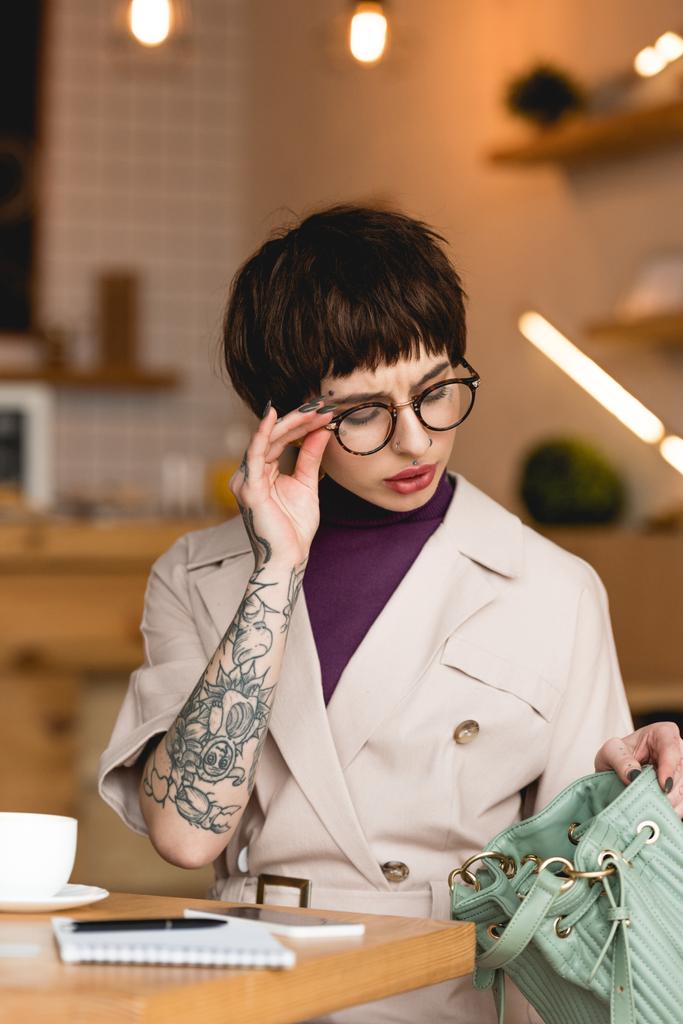 mulher de negócios atraente em óculos olhando no saco enquanto sentado no café
 - Foto, Imagem