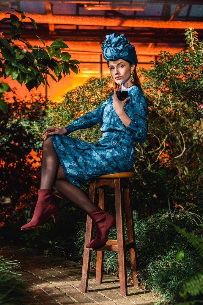 Hübsche Frau in Kleid und Turban sitzt auf Stuhl und hält Weinglas in der Orangerie - Foto, Bild