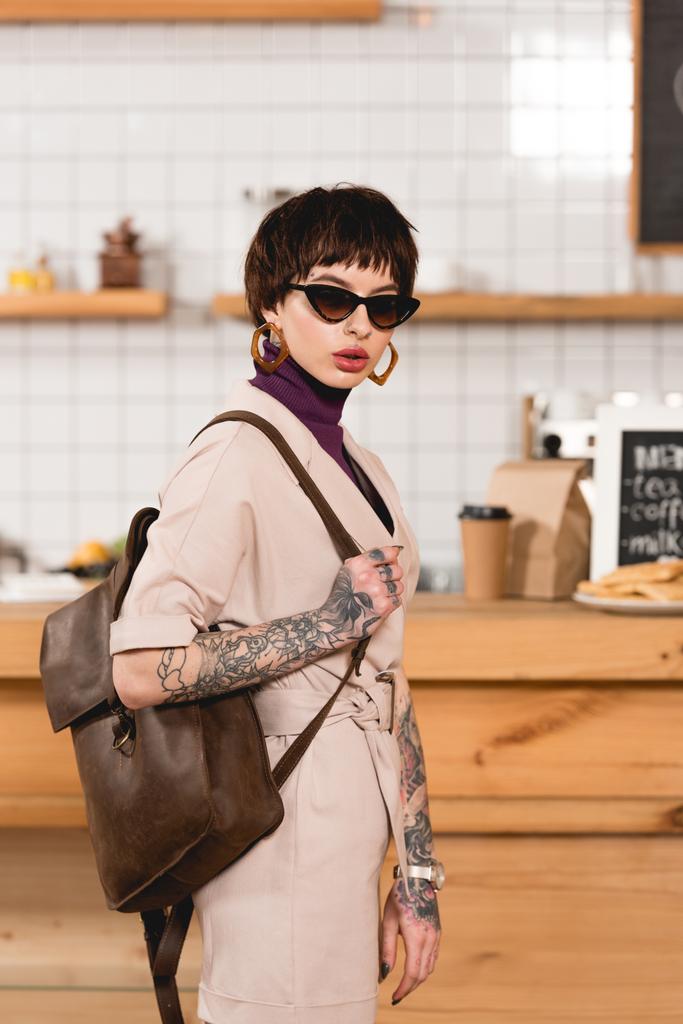 trendy, getatoeëerd zakenvrouw in zonnebril permanent in koffie winkel en camera te kijken - Foto, afbeelding