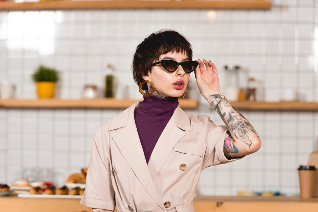 beautiful tattooed businesswoman standing in cafeteria and touching sunglasses - Photo, Image