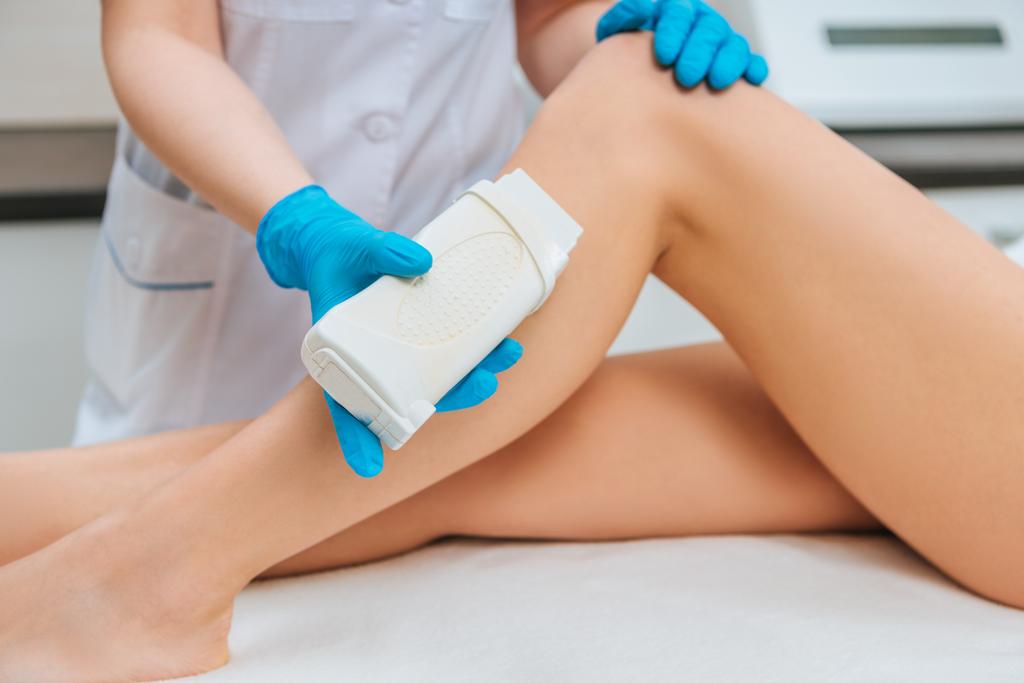 abgeschnittene Ansicht eines Kosmetikers in Gummihandschuhen, der Wachs auf das Bein aufträgt - Foto, Bild