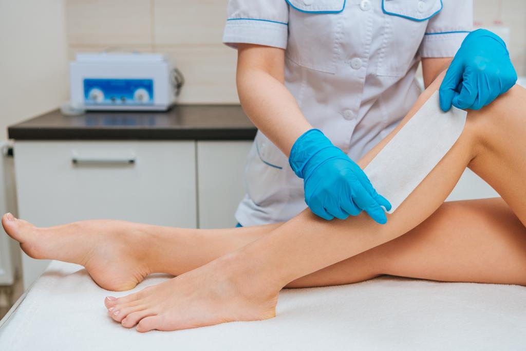 Vista parcial del cosmetólogo con guantes de goma haciendo depilación con cera de pierna
 - Foto, Imagen