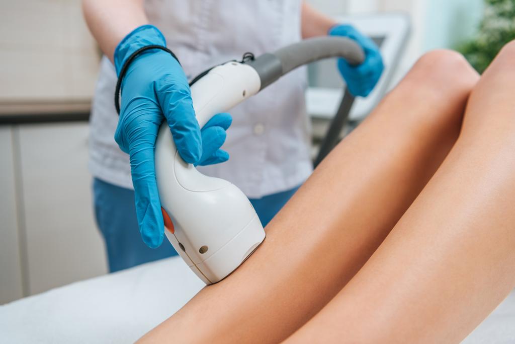 Vista parcial del cosmetólogo en guantes de goma haciendo procedimiento de depilación láser en las piernas
 - Foto, Imagen
