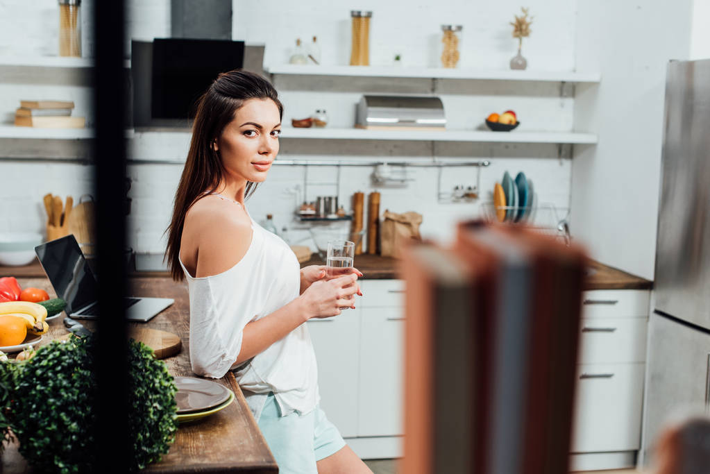 Attractive girl standing near wooden table and holding glass of water in kitchen - Photo, Image