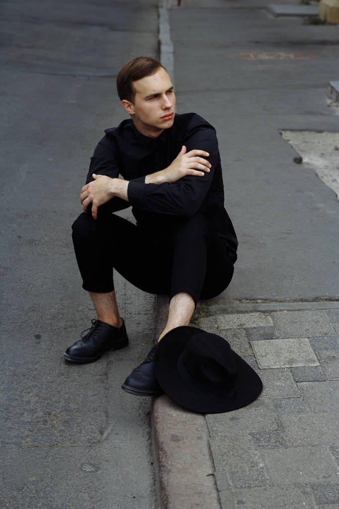 ein Mann in Schwarz mit schwarzem Hut sitzt auf dem Asphalt auf der Straße, auf der Straße. - Foto, Bild