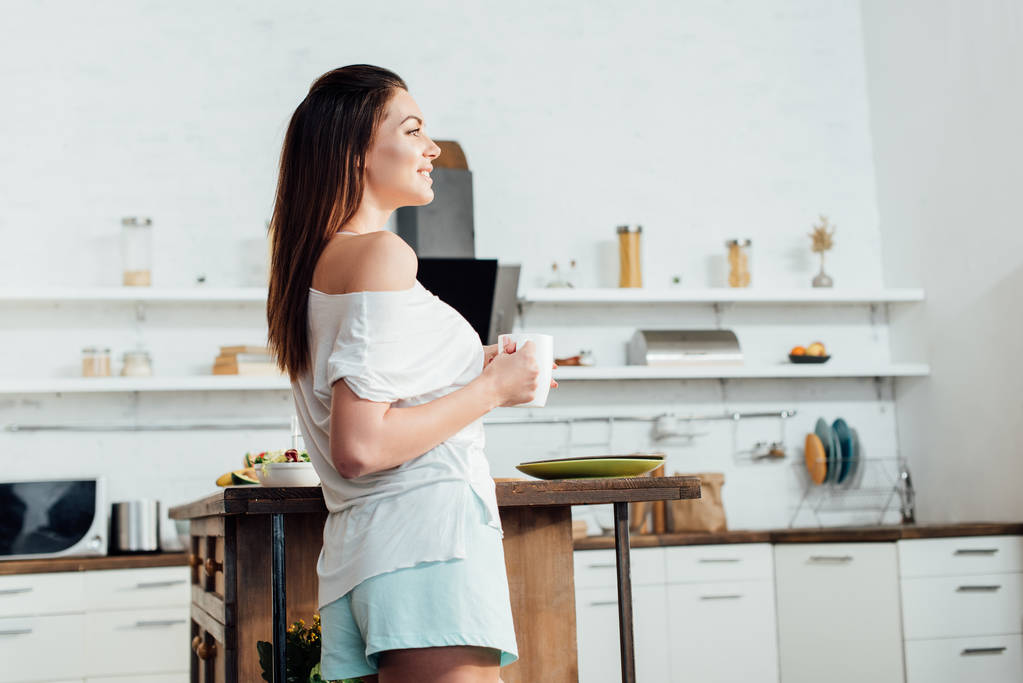 Pohled na usmívající se mladou ženu držící šálek kávy v kuchyni - Fotografie, Obrázek