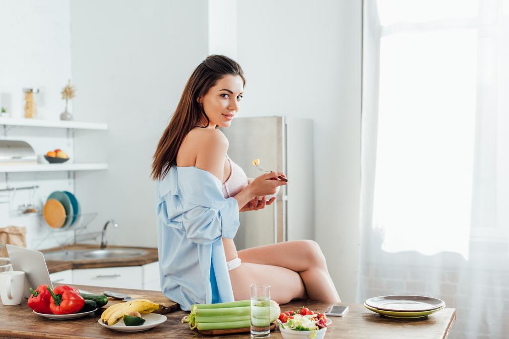 Menina sexy em roupa interior e camisa sentada na mesa e comer salada de frutas
 - Foto, Imagem